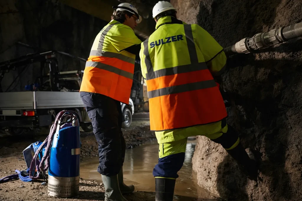 Bombas submersíveis para drenagem de obra – trabalhadores em túnel – Dois profissionais com EPIs trabalham em obra subterrânea realizando a instalação de sistema de drenagem com bomba submersível posicionada ao lado de poça d’água.
